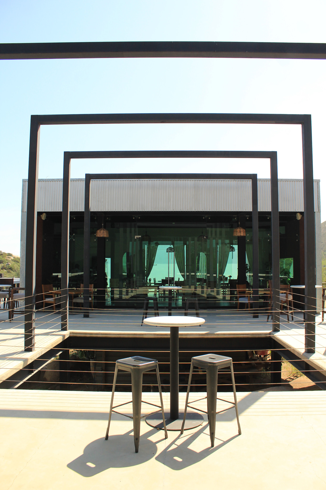 Terraza de EMAT con marcos de acero, una mesa alta y bancos frente a la fachada acristalada.