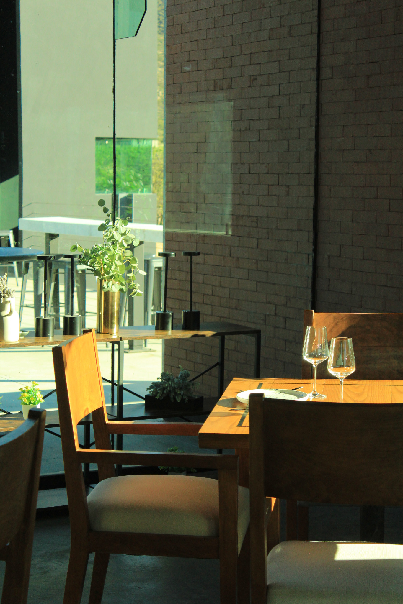 Rincón del comedor de EMAT con sillas de madera, copas, plantas y un muro de ladrillo.
