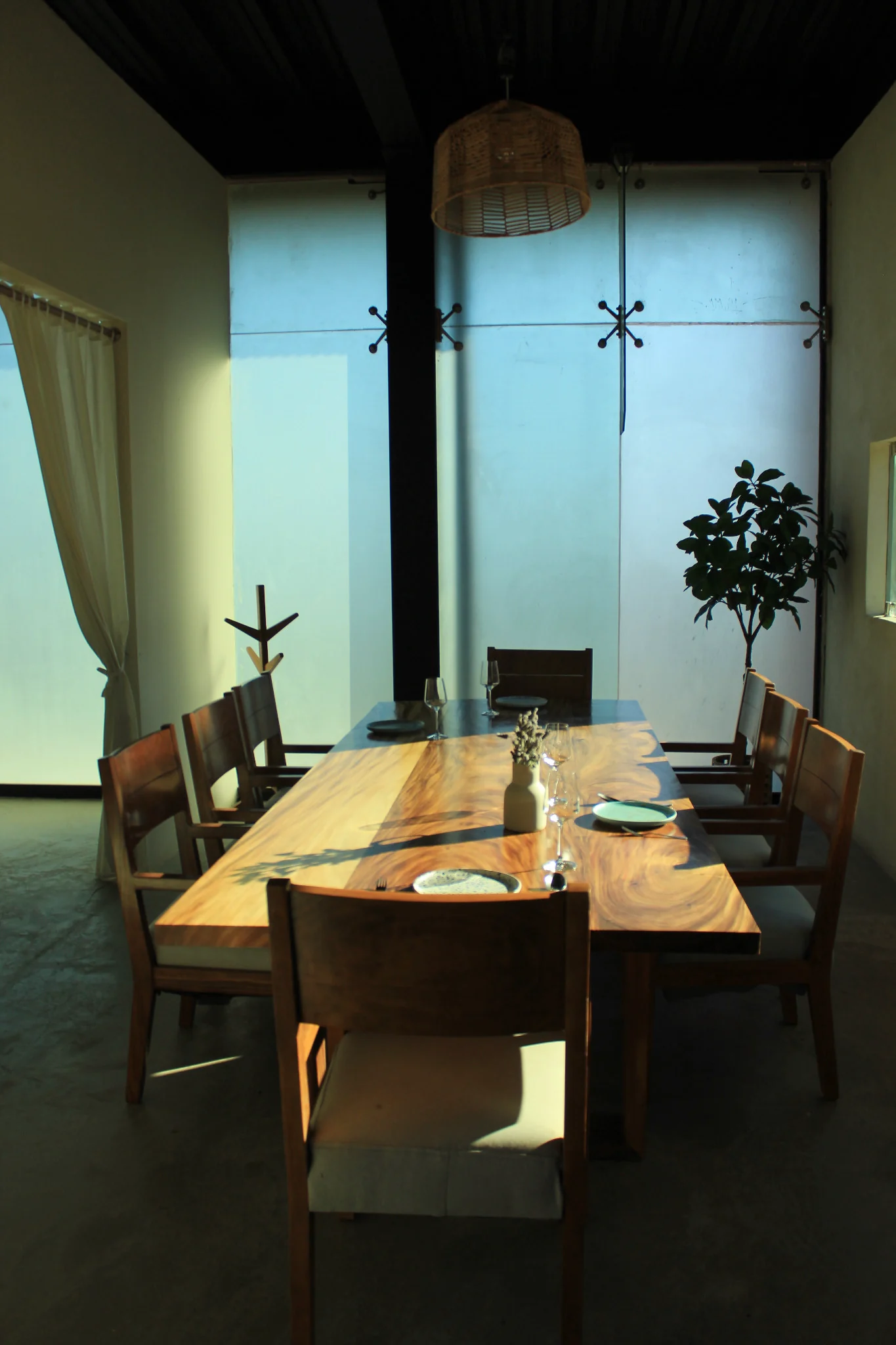 Mesa larga de madera en EMAT, con vajilla, lámpara tejida y grandes ventanales.