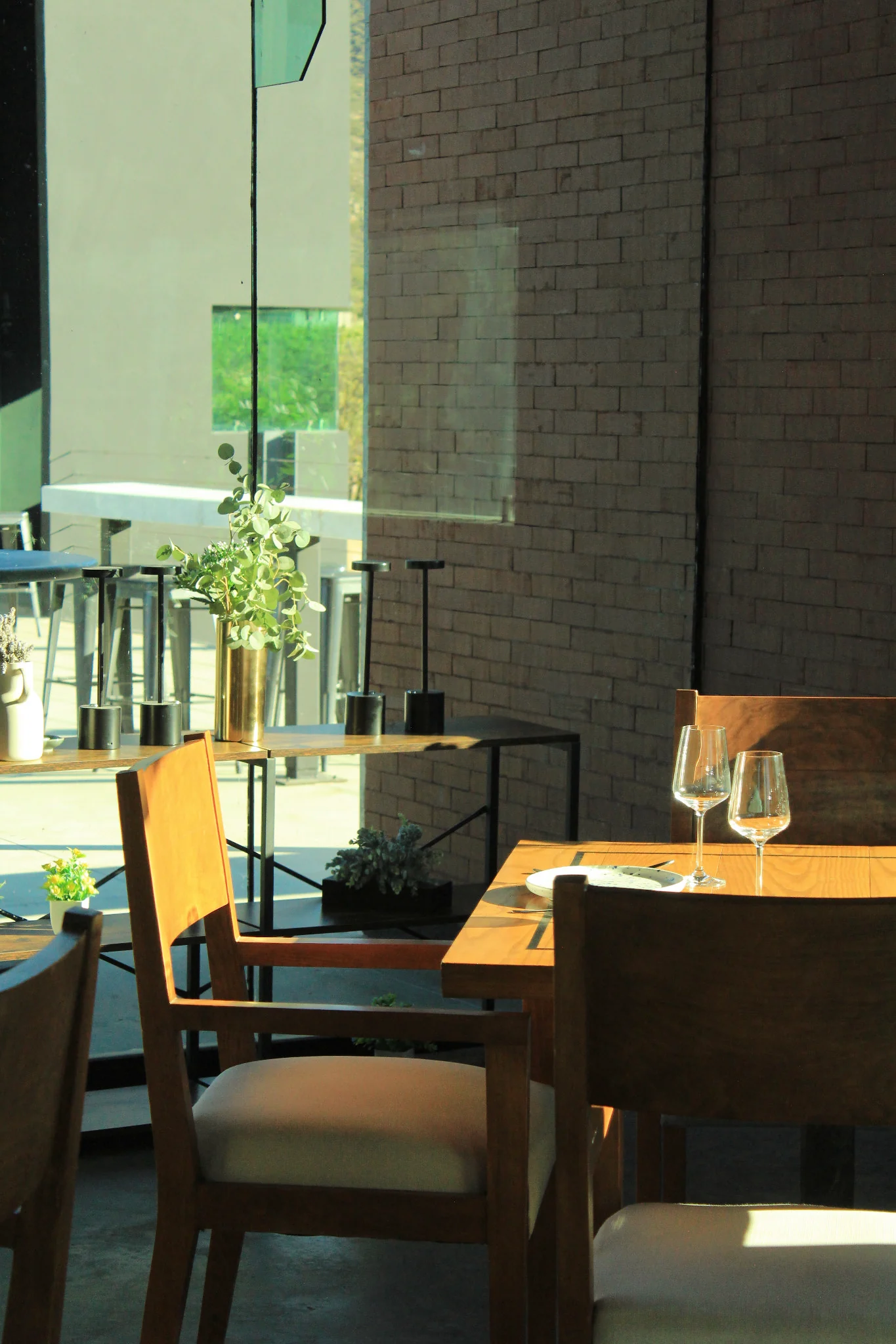 Rincón del comedor de EMAT con sillas de madera, copas, plantas y un muro de ladrillo.
